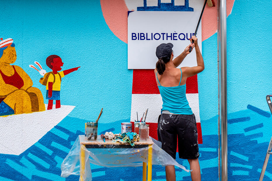 Artiste Élice peint une fresque sur une façade de bibliothèque, représentant un océan de livres avec un groupe de personnage dans un papier en origami